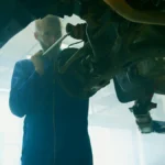 Mekanik berpakaian biru melakukan perawatan kendaraan di bengkel, menggambarkan arti maintain